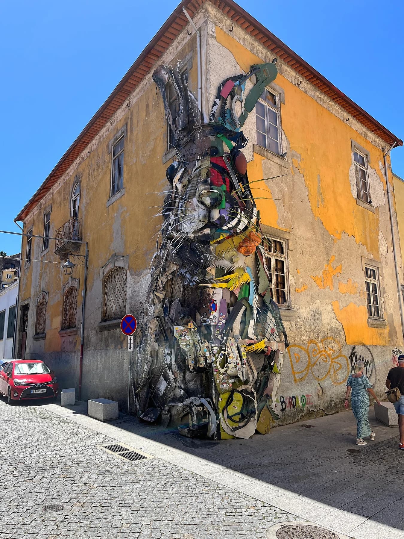 Bordalo II’s large Half Rabbit sculpture made from discarded materials extending around the corner of a yellow building in Gaia.