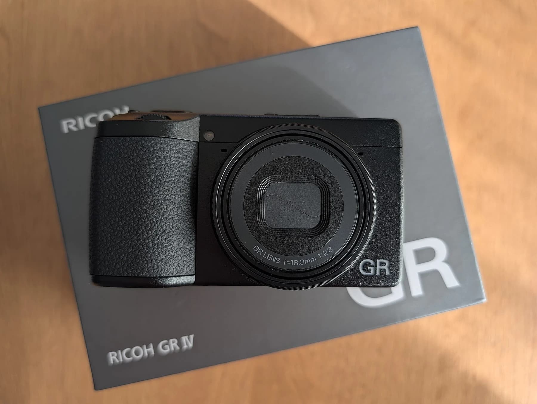 A black Ricoh GR IV compact camera resting on its original box on a warm wooden surface, seen from the front.