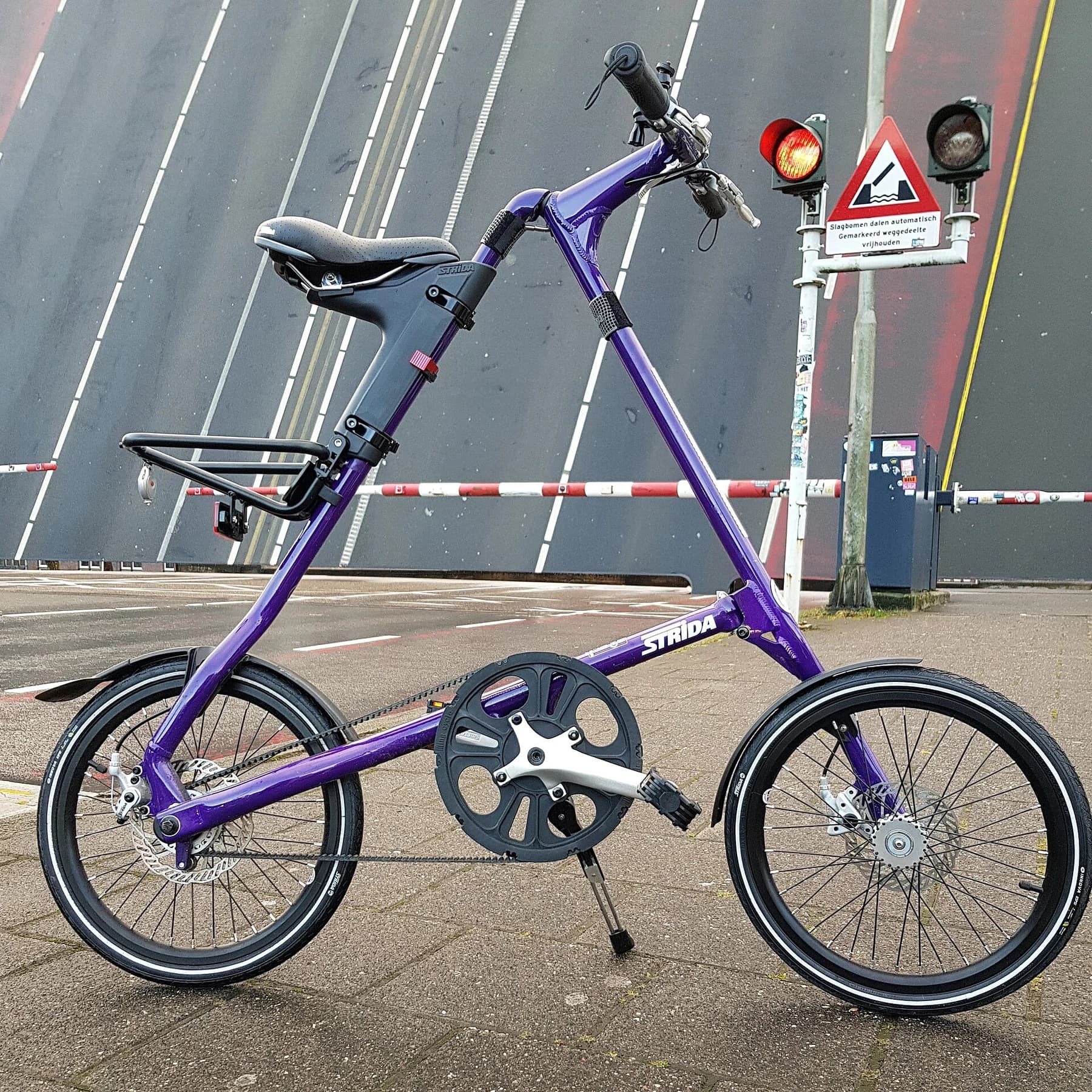 A purple Strida SX bicycle in side profile, showing its triangular frame, belt drive and small wheels beside a raised bridge.