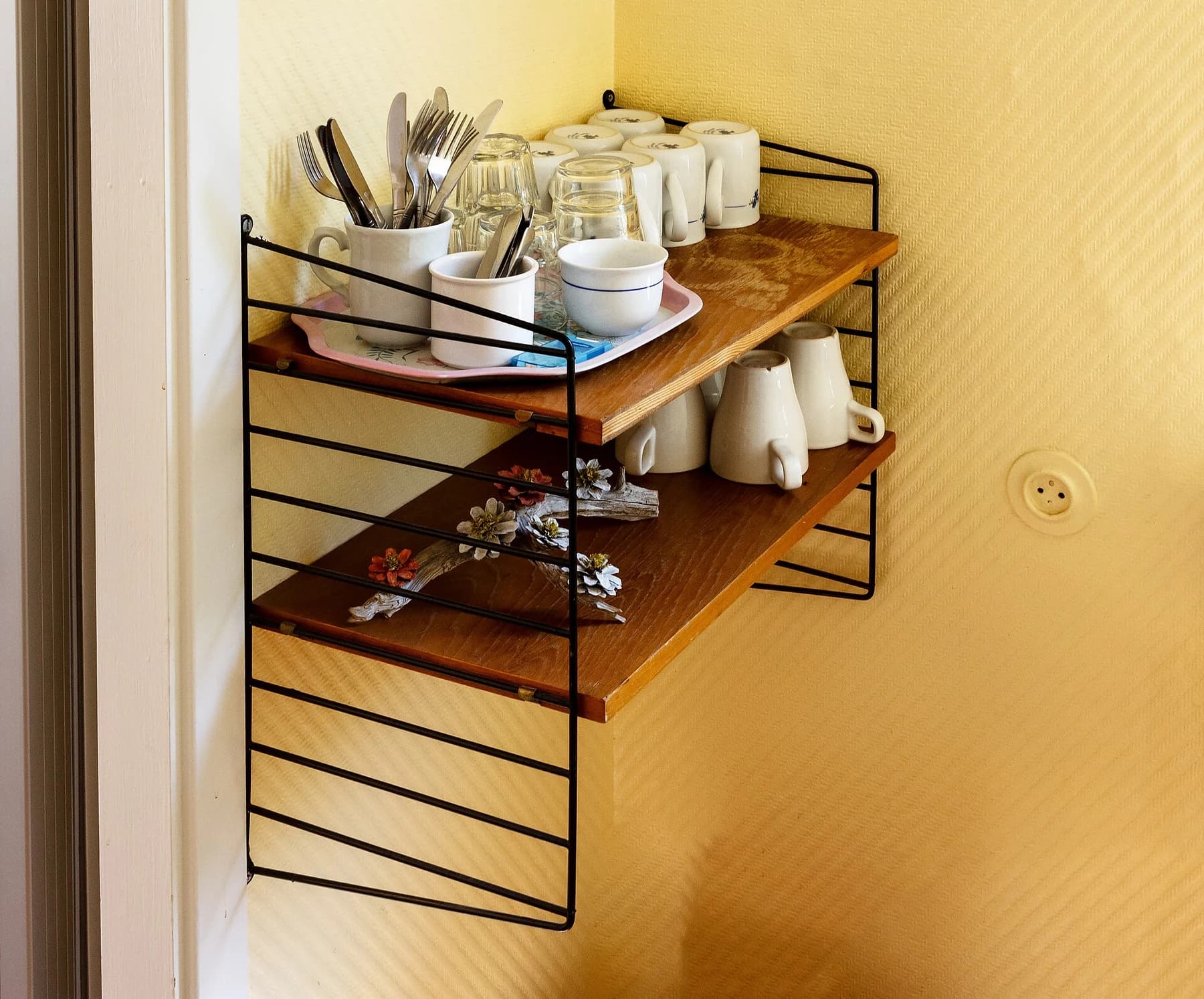 Black wire String shelving holding cups, glasses and cutlery against a sunlit yellow wall in a Swedish bakery.