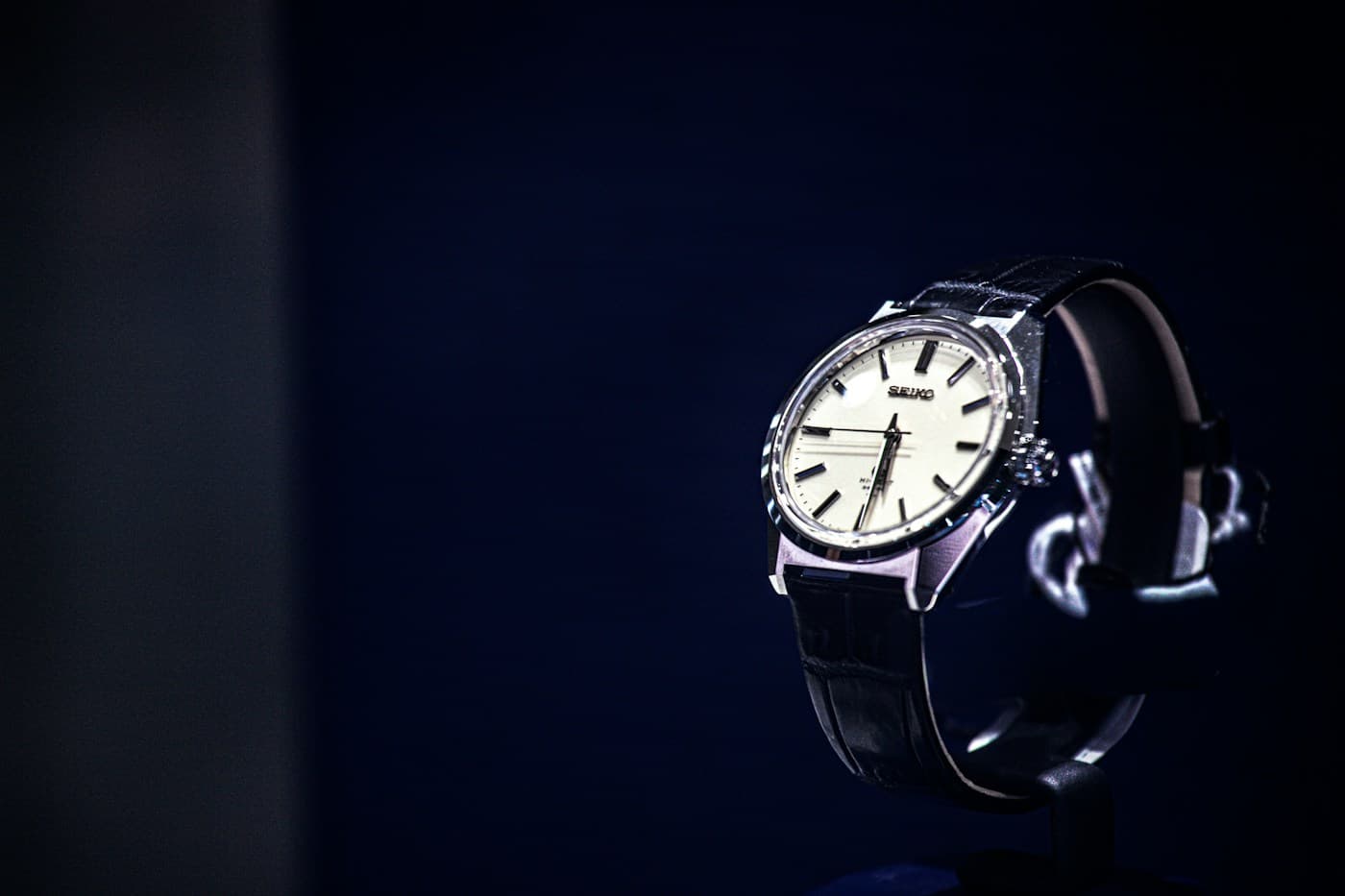 A silver-coloured wristwatch with a black leather strap, photographed against a deep blue background.